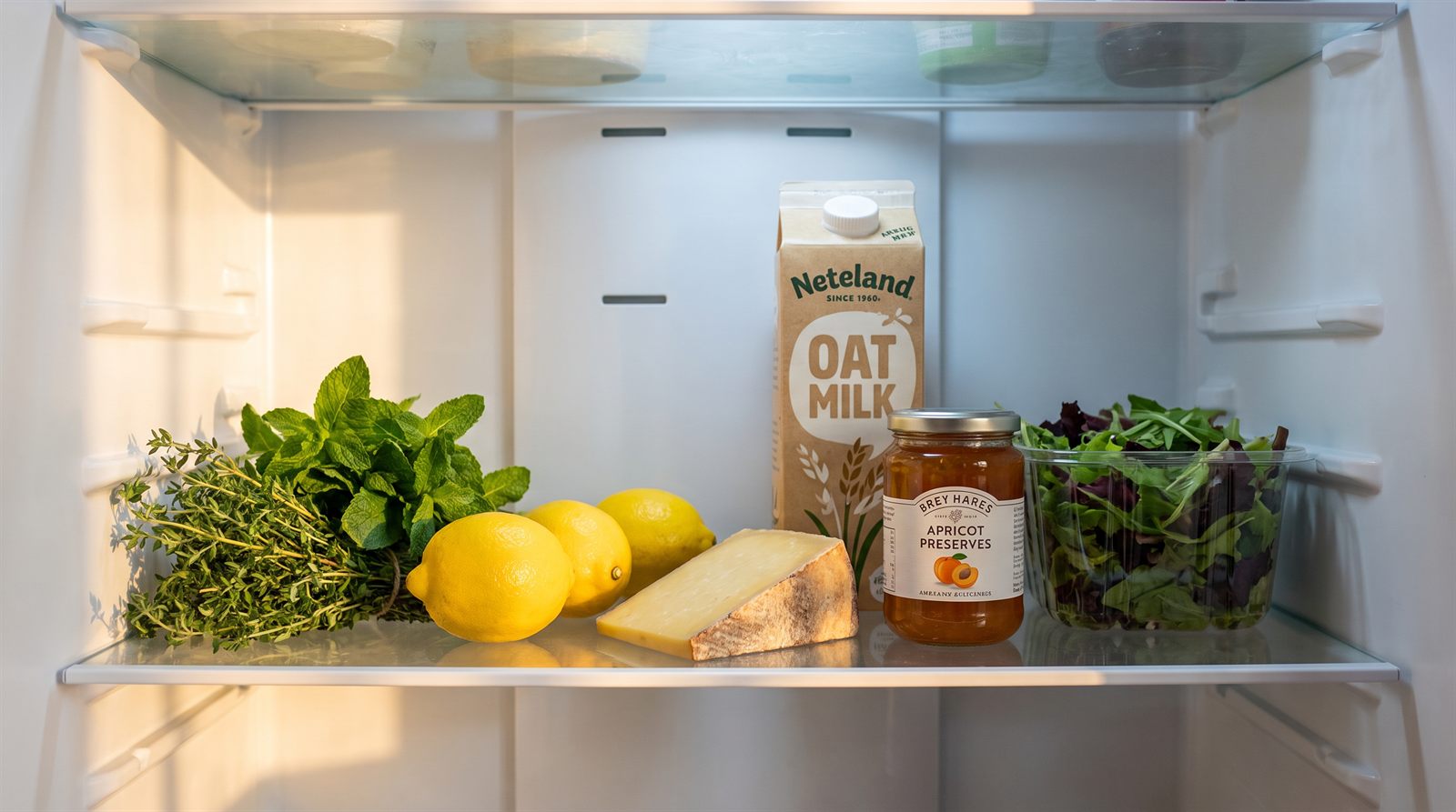 One refrigerator shelf holding herbs, lemons, cheese, a carton, a jar and salad leaves, each clearly separate